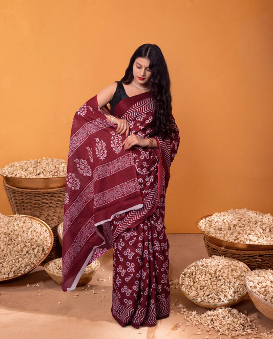 A woman models a deep maroon bandhani saree with white floral and geometric prints, paired with a black sleeveless blouse, set against a warm orange background with woven baskets of rice.