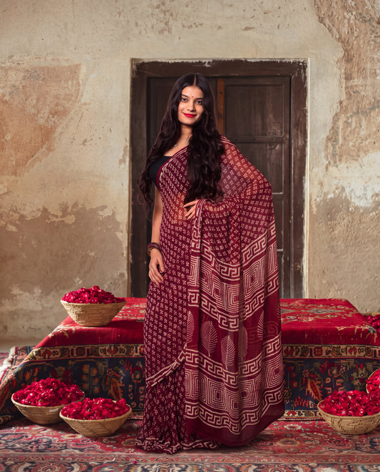 A woman wearing a deep maroon and burgundy saree with geometric patterns stands in front of a rustic wall and wooden door. Baskets of red rose petals are scattered around her on patterned rugs.