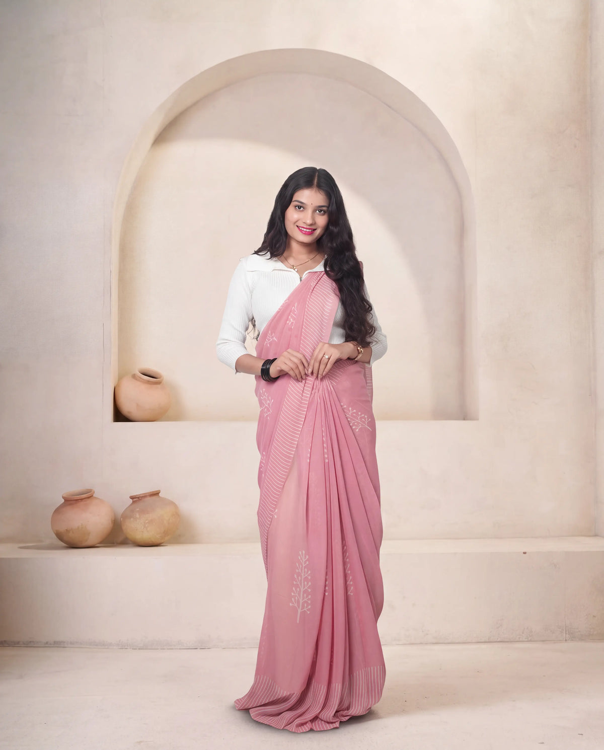 A woman modeling a pink saree with white embroidery, paired with a white long-sleeve blouse, standing in front of a beige plaster archway with decorative clay pots.