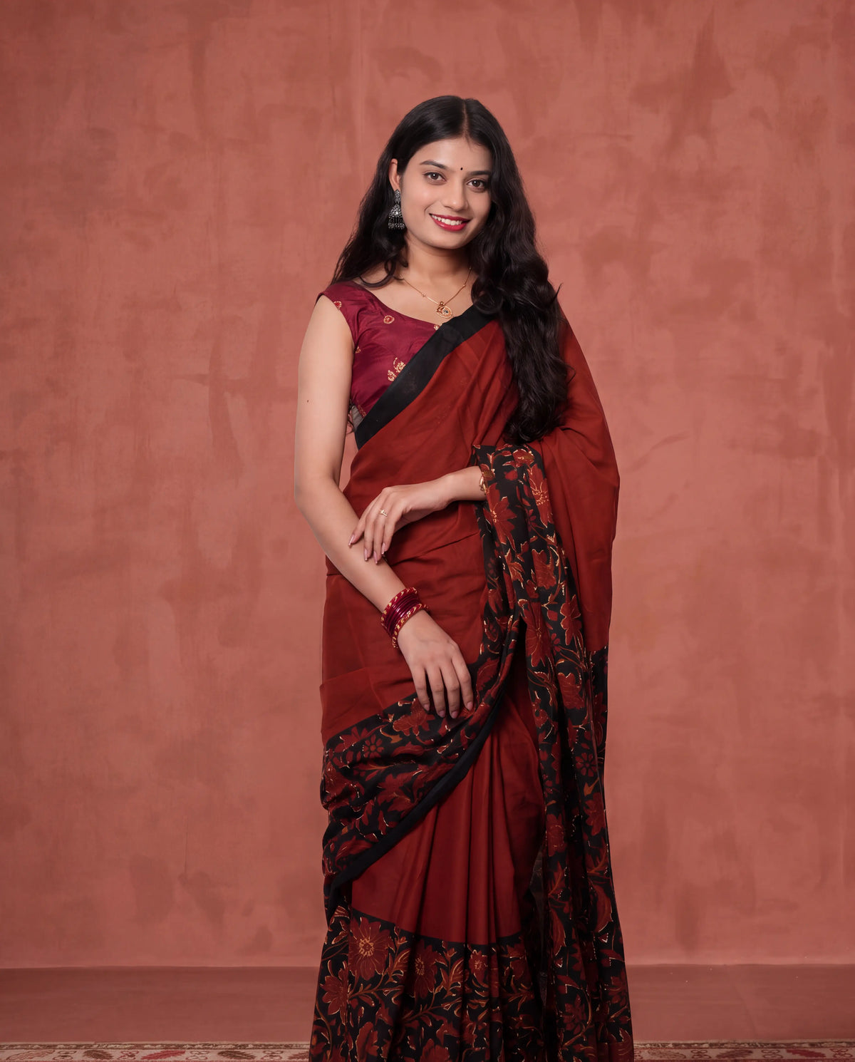 A woman modeling a deep maroon red saree with black borders and a black floral patterned pallu, paired with a matching maroon blouse, set against a textured terracotta wall.