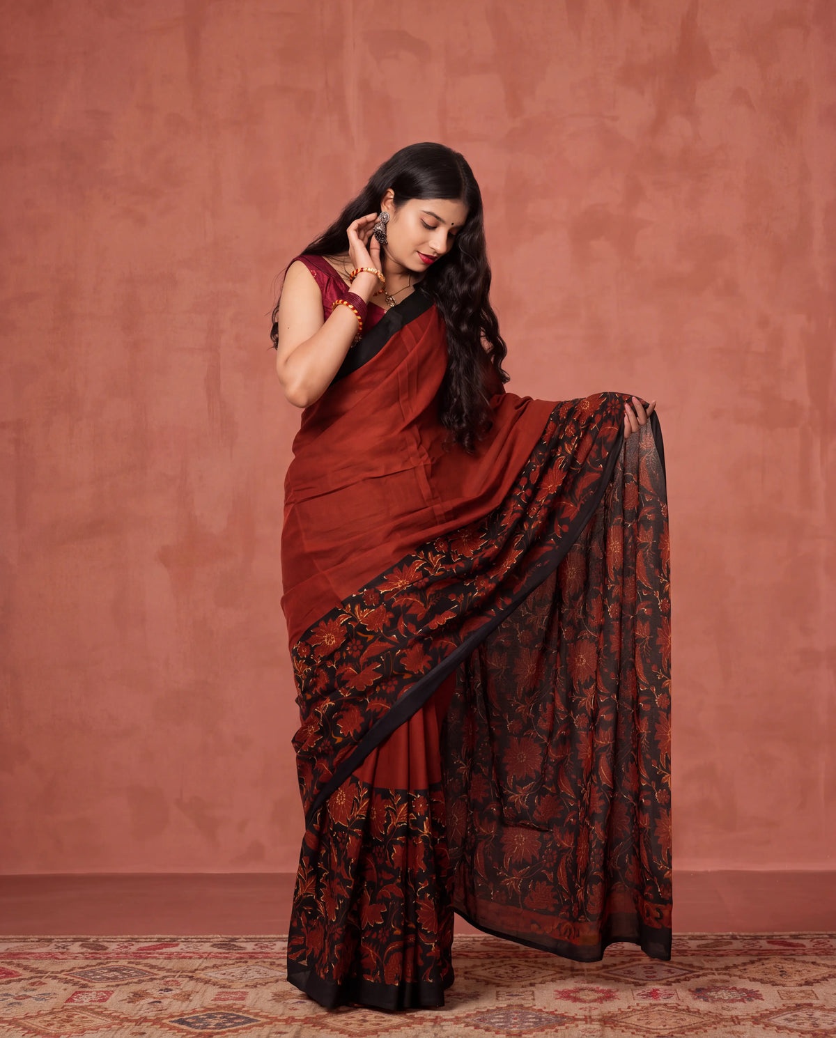 A woman models a dark red and black saree with floral embroidery, standing against a textured terracotta-colored wall.
