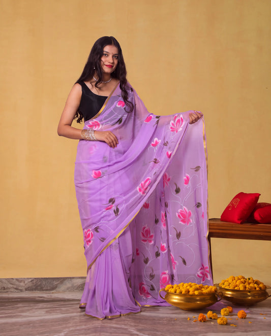 A woman models a lavender purple saree with pink floral prints and a black sleeveless blouse, standing against a warm beige wall with decorative props.
