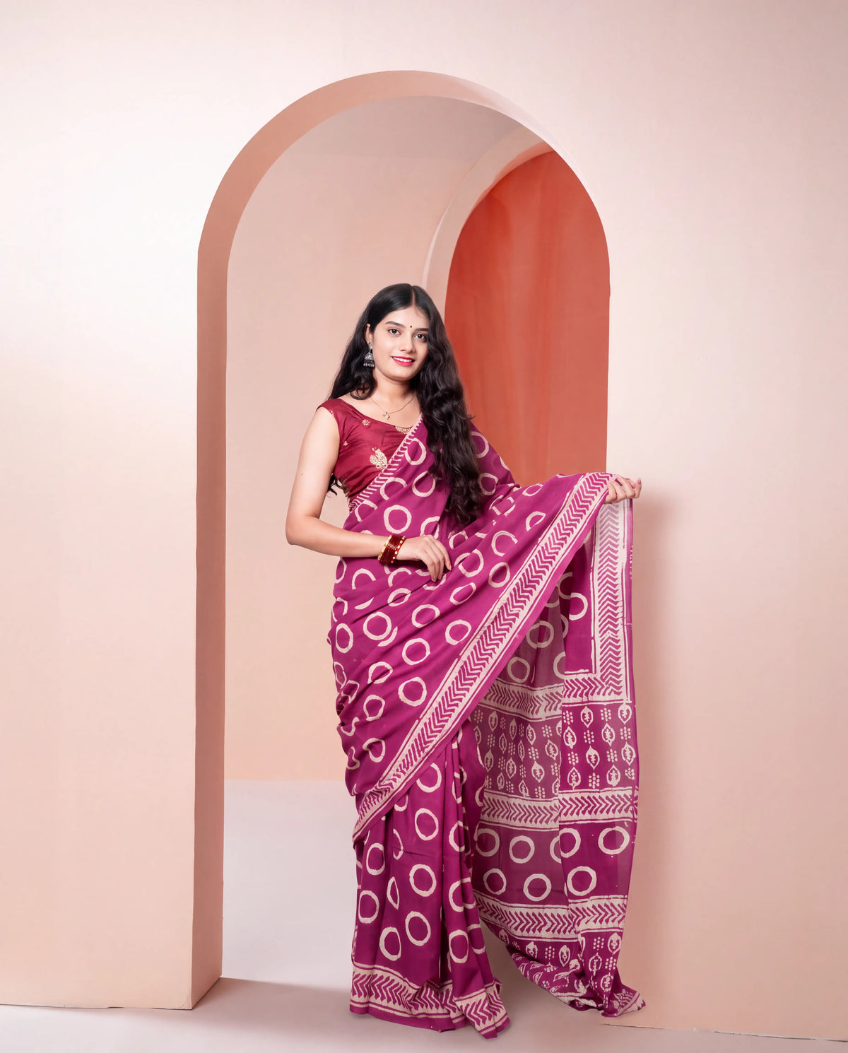 A woman models a magenta purple saree with white circular and geometric patterns, paired with a matching sleeveless blouse, standing in front of a peach-colored arched wall.