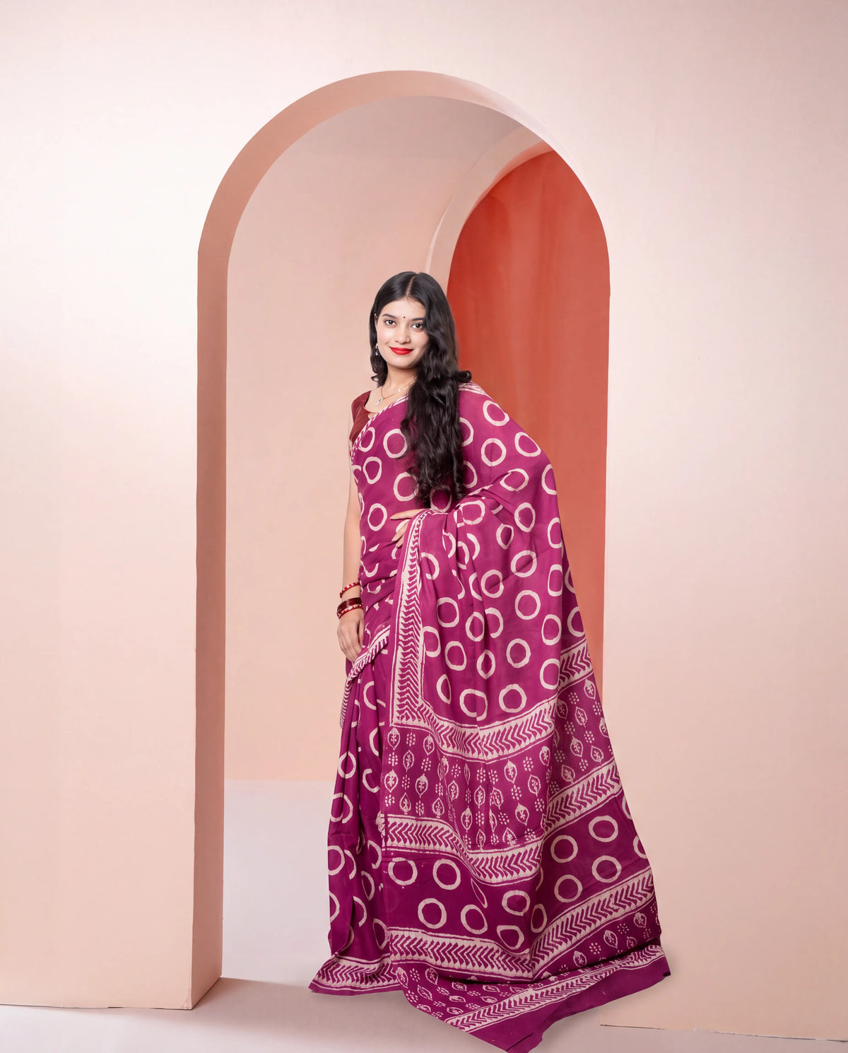 A woman modeling a magenta purple saree with white bandhani circle and geometric patterns, standing in front of a peach-colored arched doorway.