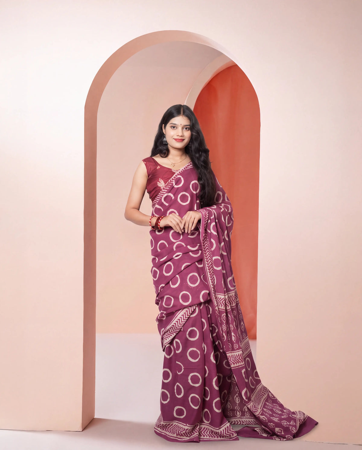 A woman modeling a purple saree with a white circular pattern and a matching purple blouse, standing in front of a peach-colored arched backdrop.