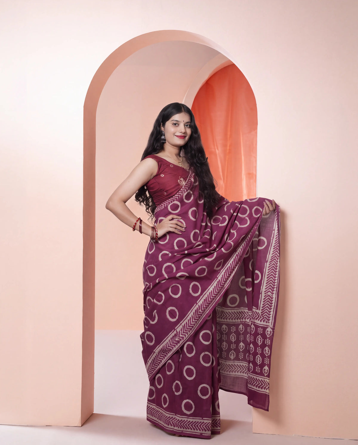 A woman models a deep purple saree with white circular patterns and a matching sleeveless blouse, standing in a peach-colored arched doorway.
