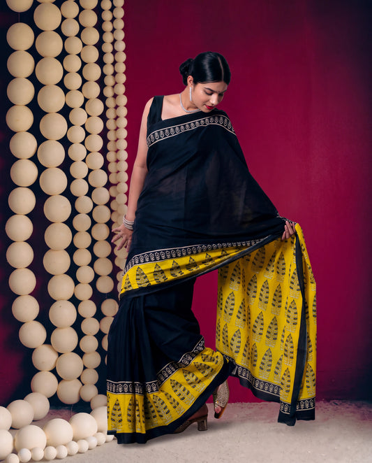 A woman models a black and yellow saree with intricate border patterns, posed in front of a dark red backdrop and a decorative wall of beige spheres.