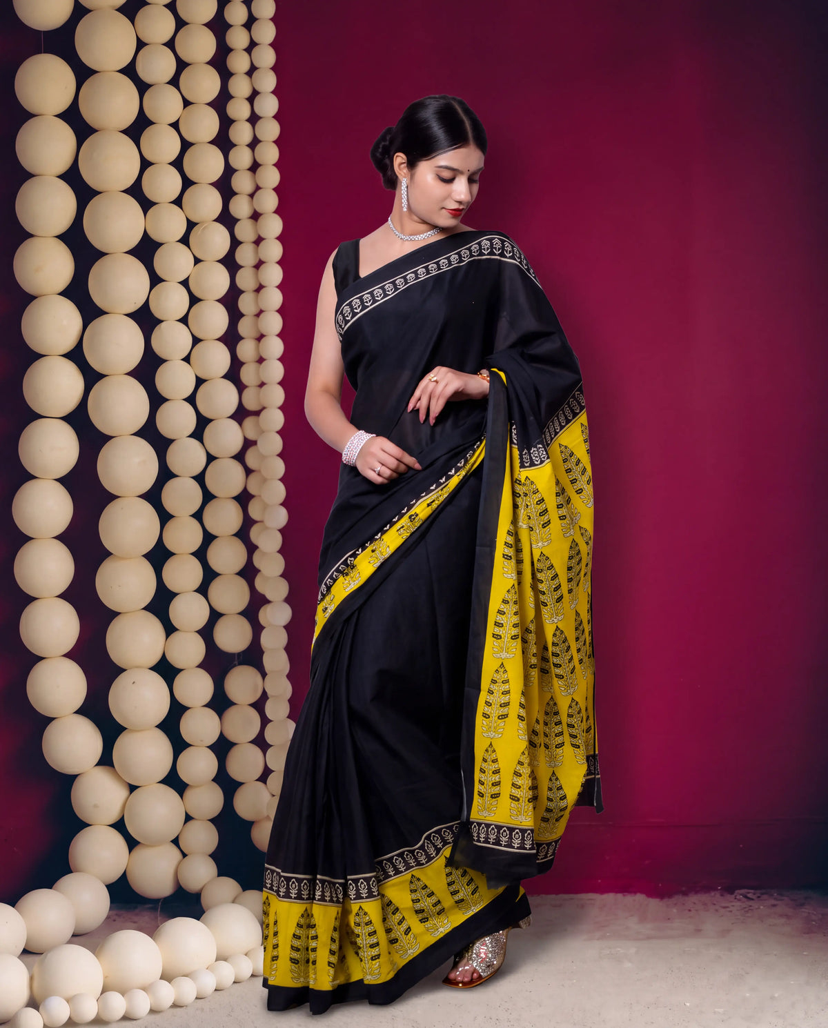 A woman modeling a black and yellow saree with intricate black patterns on the border and pallu, standing against a deep magenta backdrop with a decorative wall of hanging beige spheres on the left.