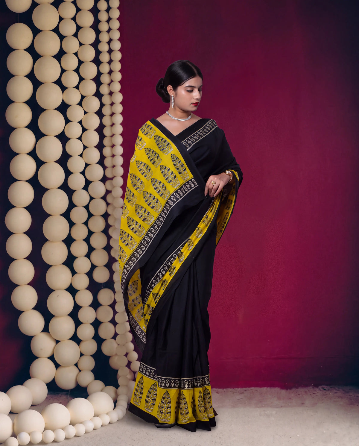 A woman models a black and mustard yellow saree with intricate geometric borders, standing in front of a dark magenta wall decorated with hanging beige spheres.