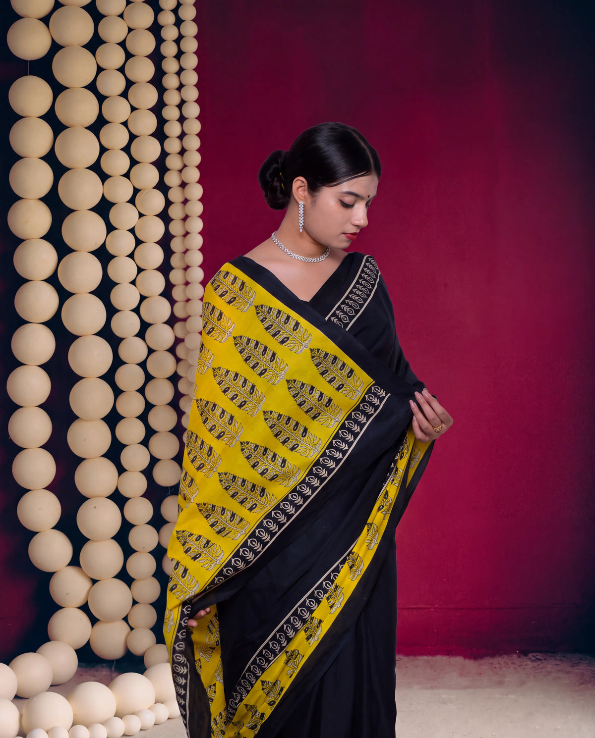 A woman wearing a black and yellow saree with intricate gold embroidery, standing in front of a dark red wall with a decorative column of beige spheres on the left.