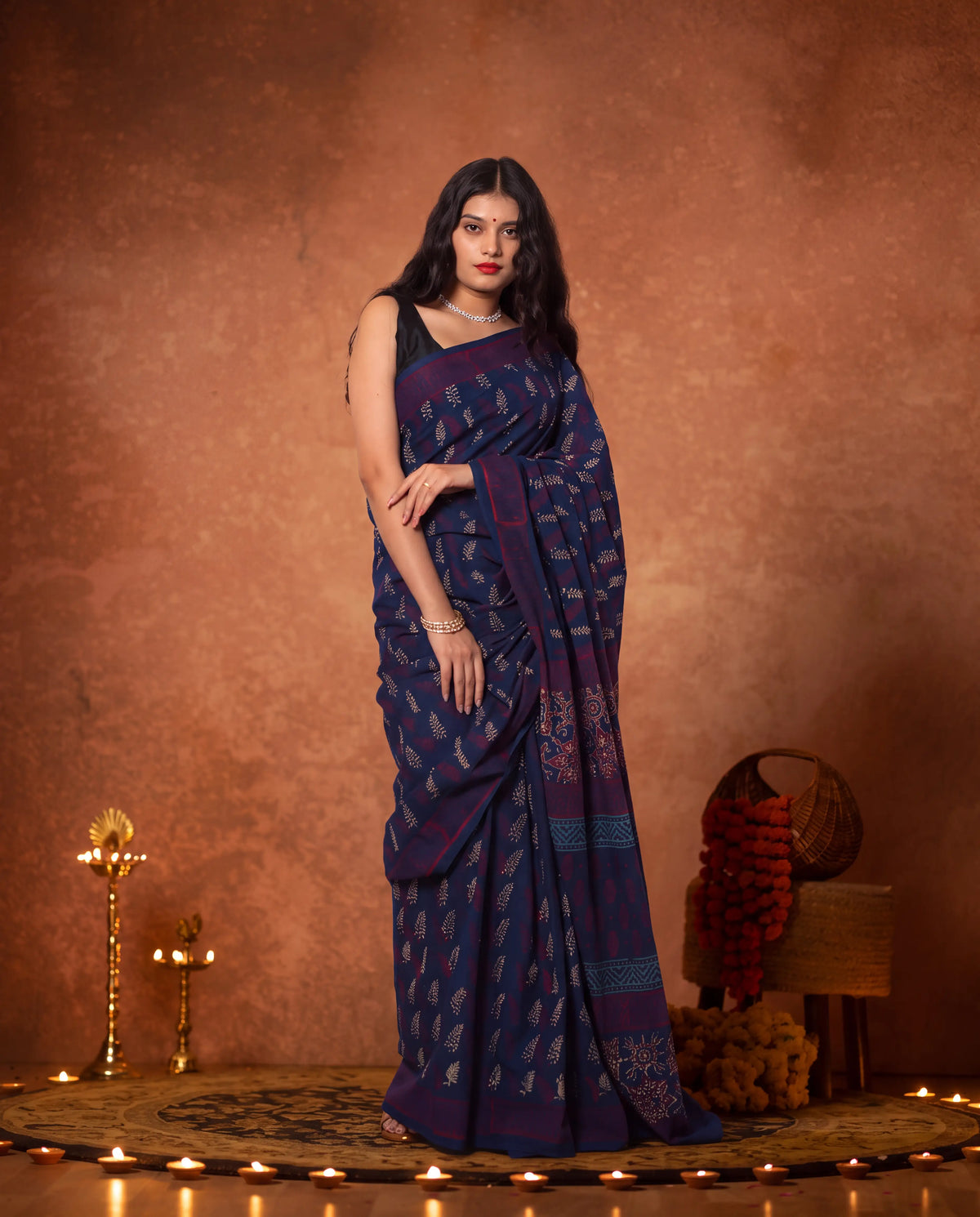 A woman models a dark navy blue saree with intricate gold and silver floral patterns, standing against a textured warm brown backdrop decorated with traditional Indian lamps and a basket.