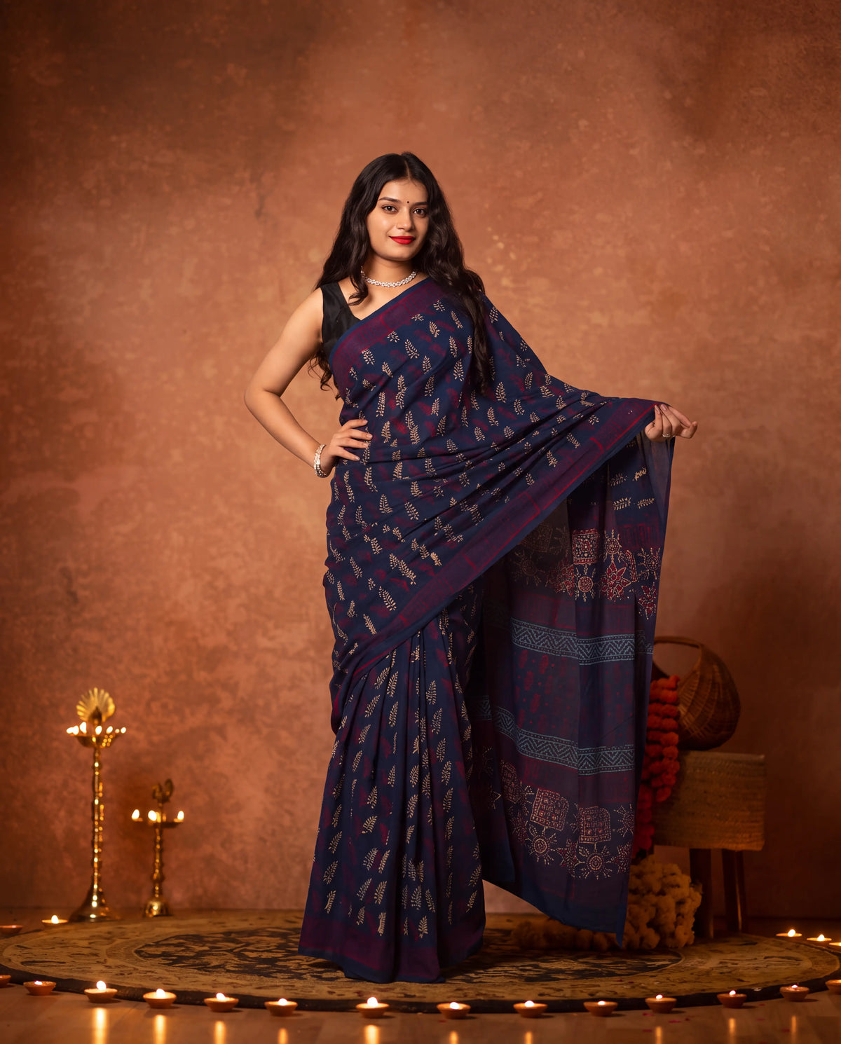 A woman models a dark navy blue saree with intricate gold and silver floral patterns, posed against a textured warm brown backdrop with decorative candles and traditional props.