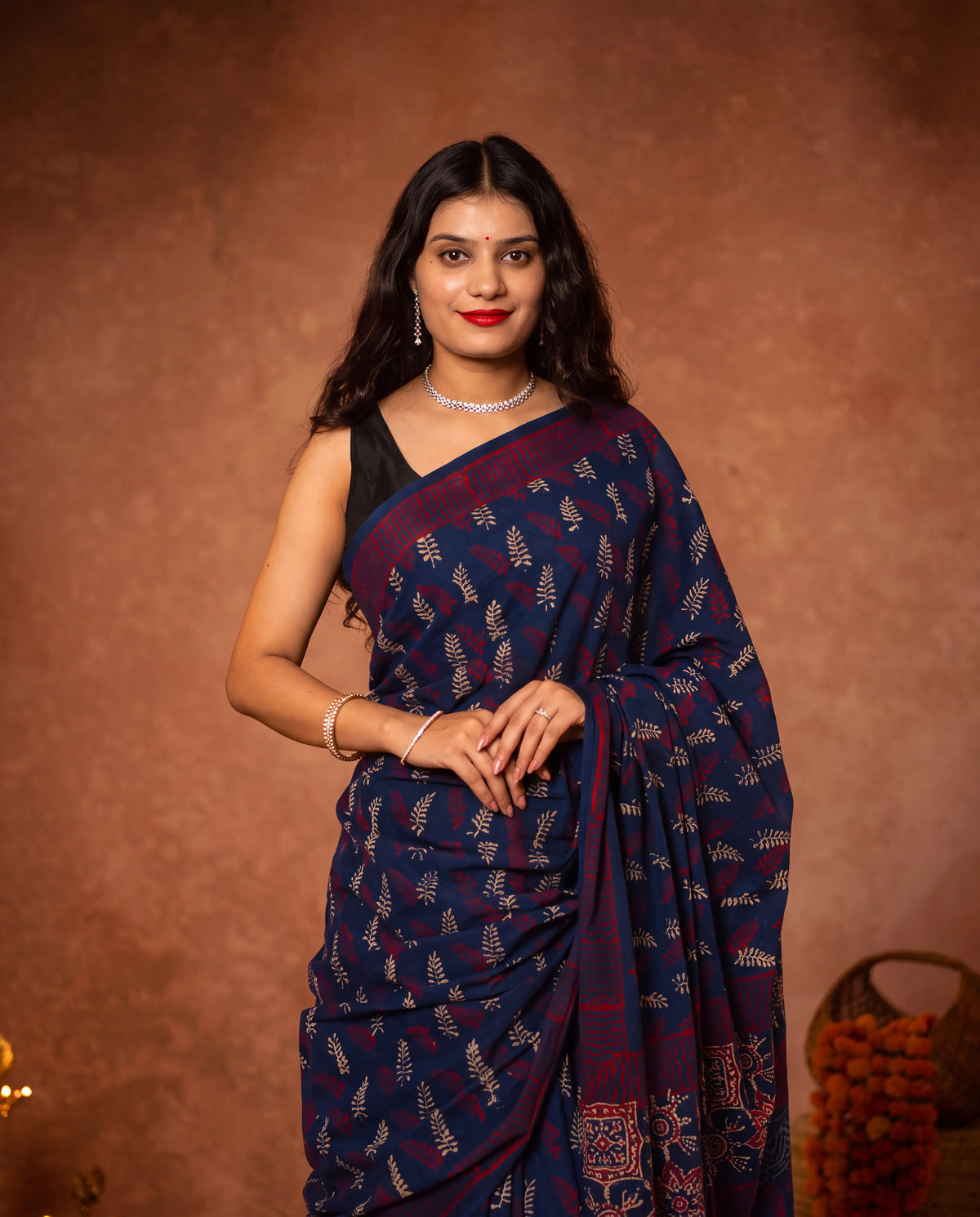 A woman wearing a navy blue saree with red and gold block prints and a black sleeveless blouse, standing against a textured brown studio backdrop.