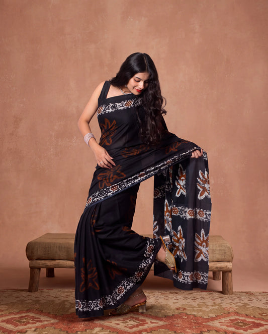 A woman models a black saree with brown and white floral patterns, sitting on a rustic wooden bench against a textured terracotta background.