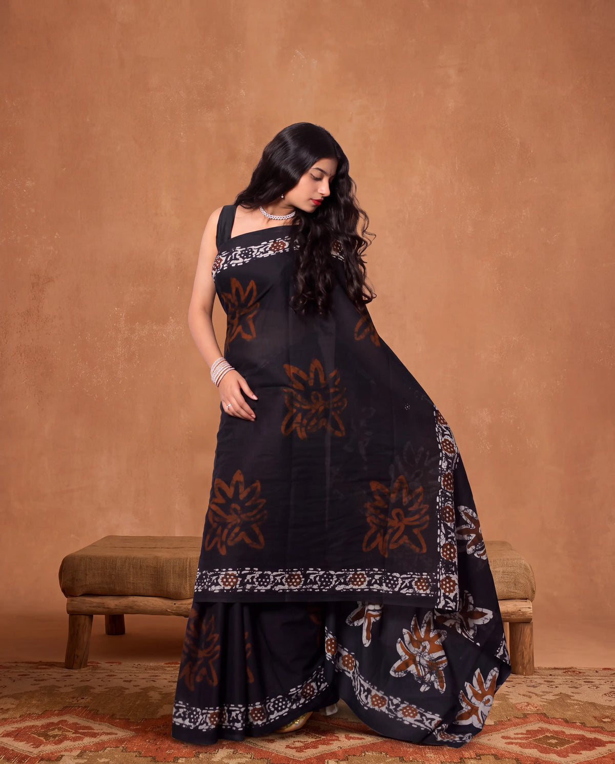 A woman models a black georgette saree with brown and white floral block prints and borders, paired with matching black palazzo pants, set against a warm brown textured backdrop.