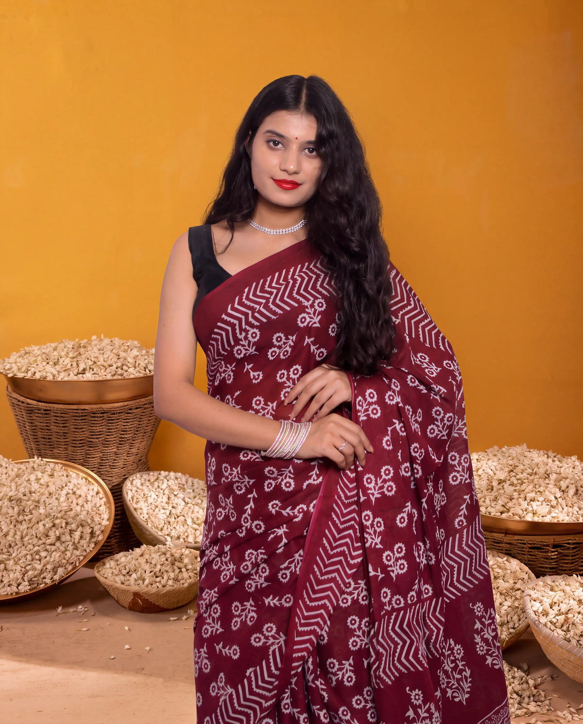 A woman modeling a deep maroon saree with white bandhani patterns, paired with a black sleeveless blouse, standing in front of a warm yellow background with woven baskets of rice.
