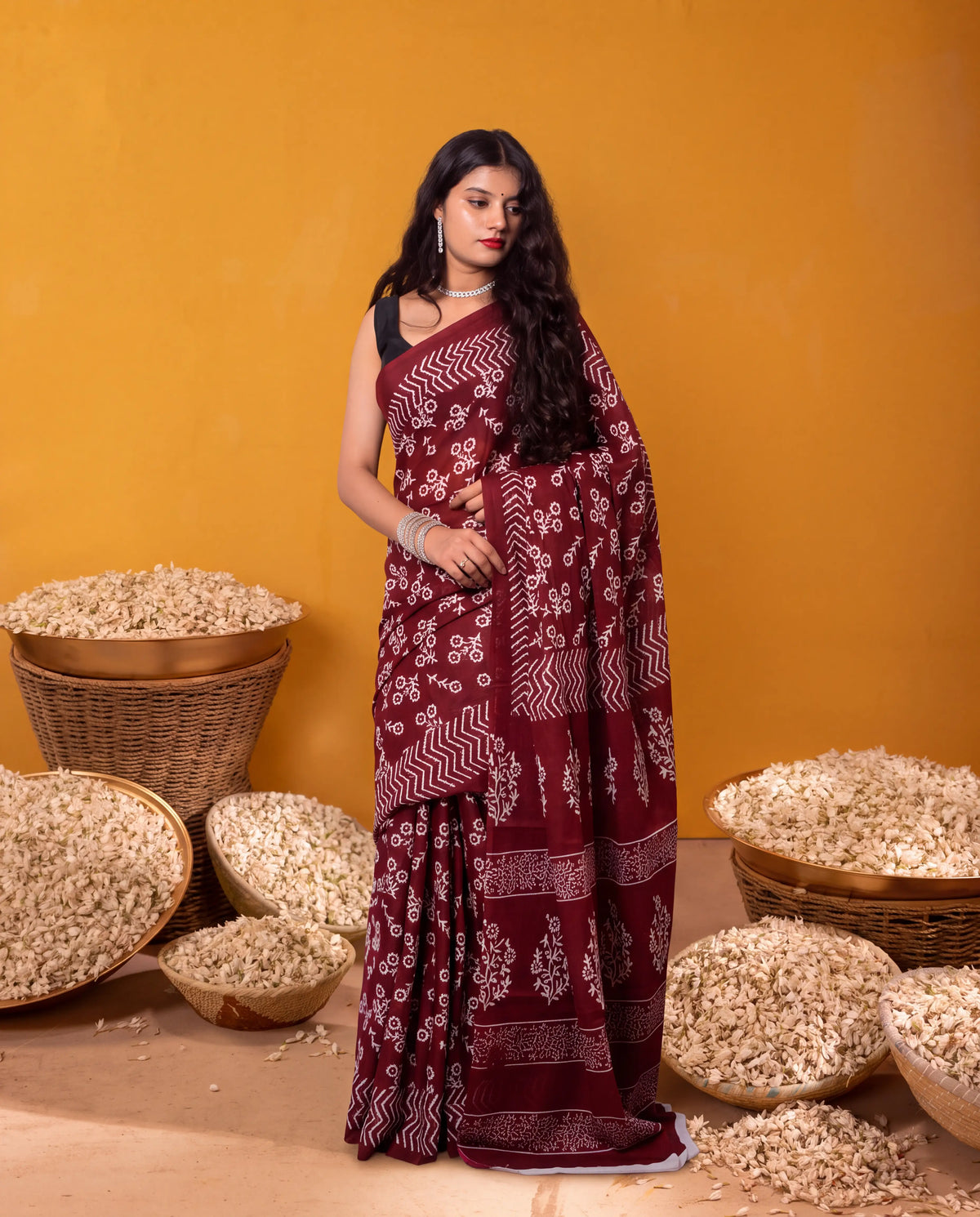 A woman modeling a deep maroon saree with white block prints, standing against a warm yellow-orange background with woven baskets of rice scattered around her.