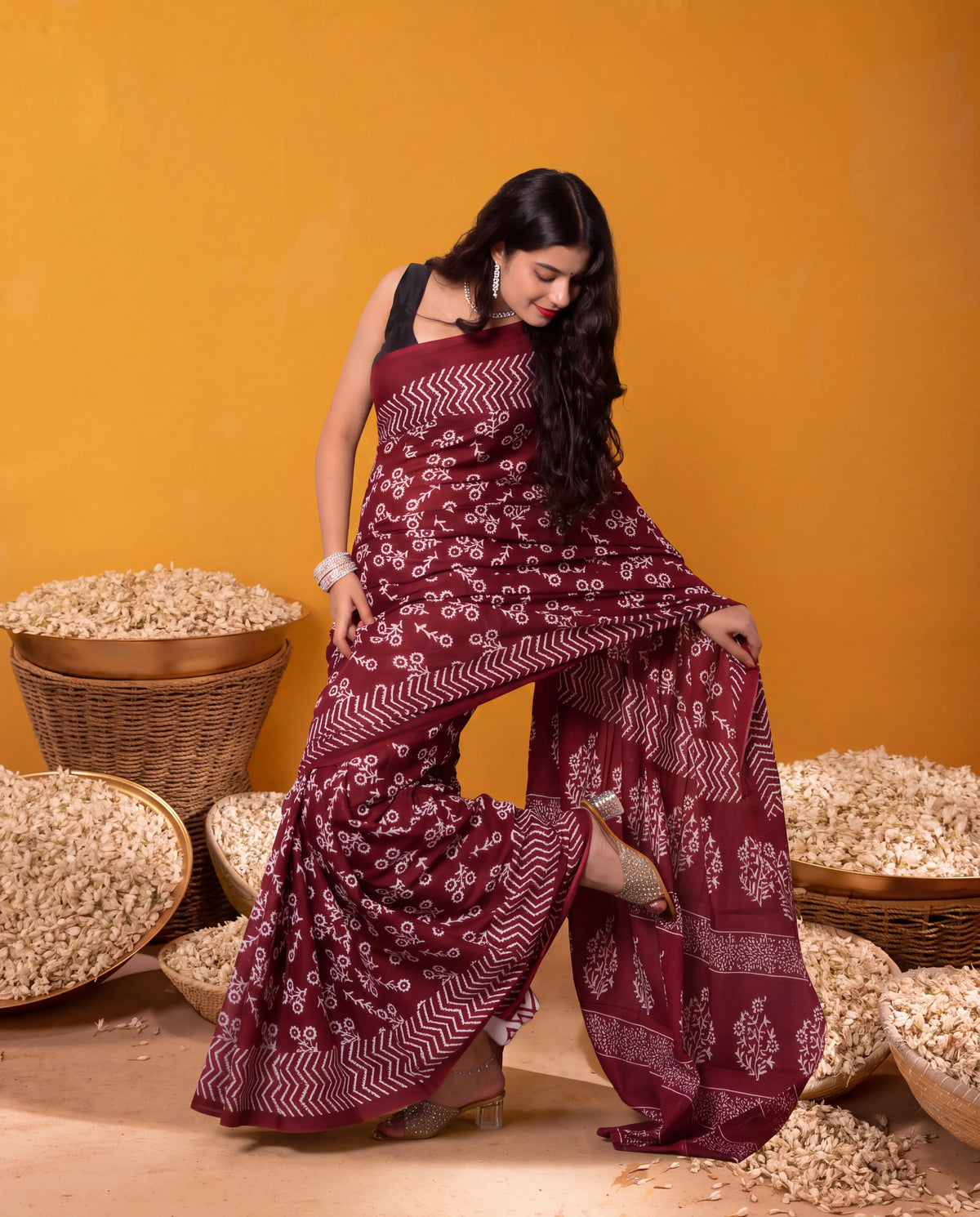 A woman models a deep maroon saree with white block prints and chevron patterns, posed in front of a warm yellow-orange background with woven baskets of dried flowers.