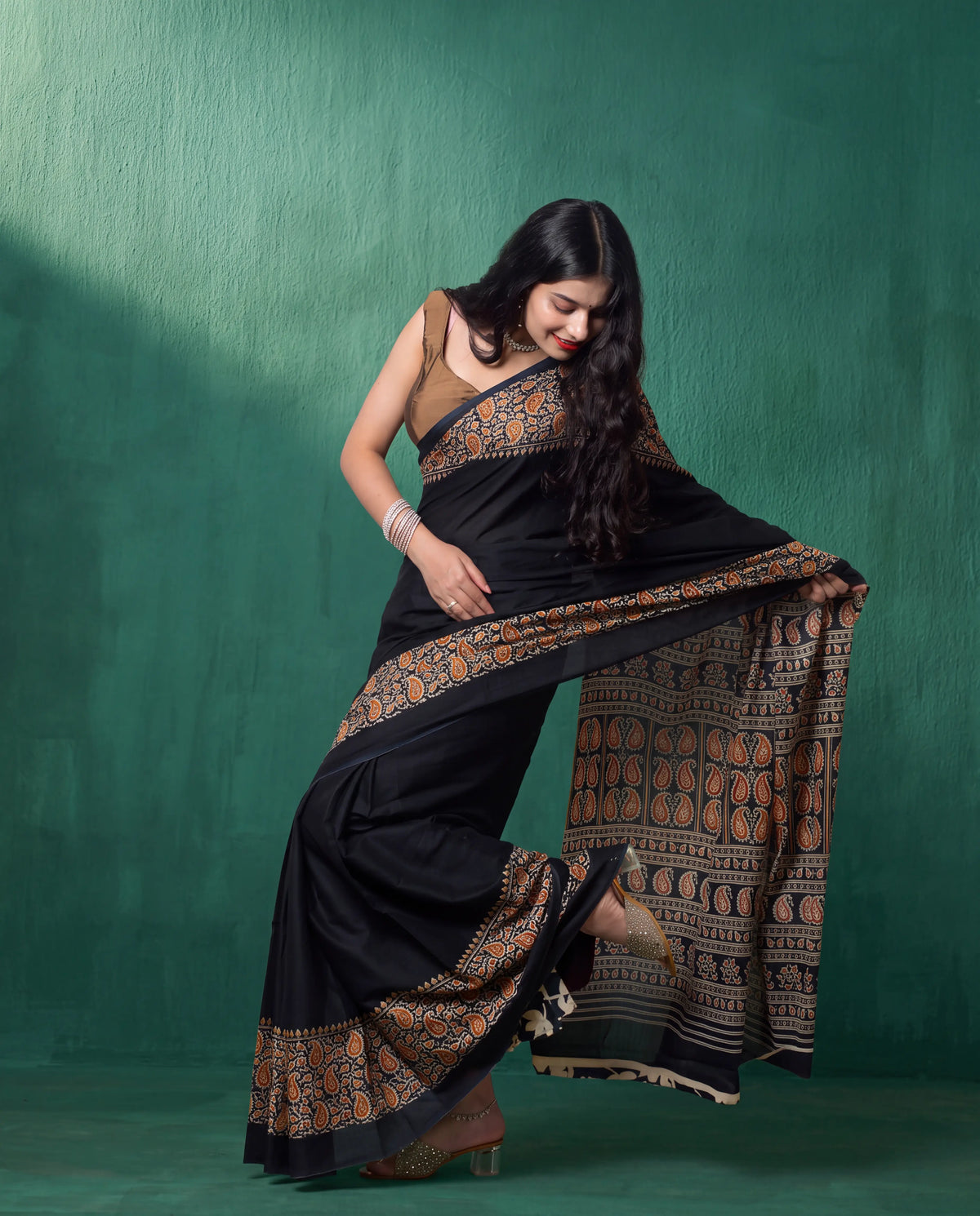 A woman modeling a black saree with intricate gold and brown border patterns, posed against a textured dark green studio backdrop.