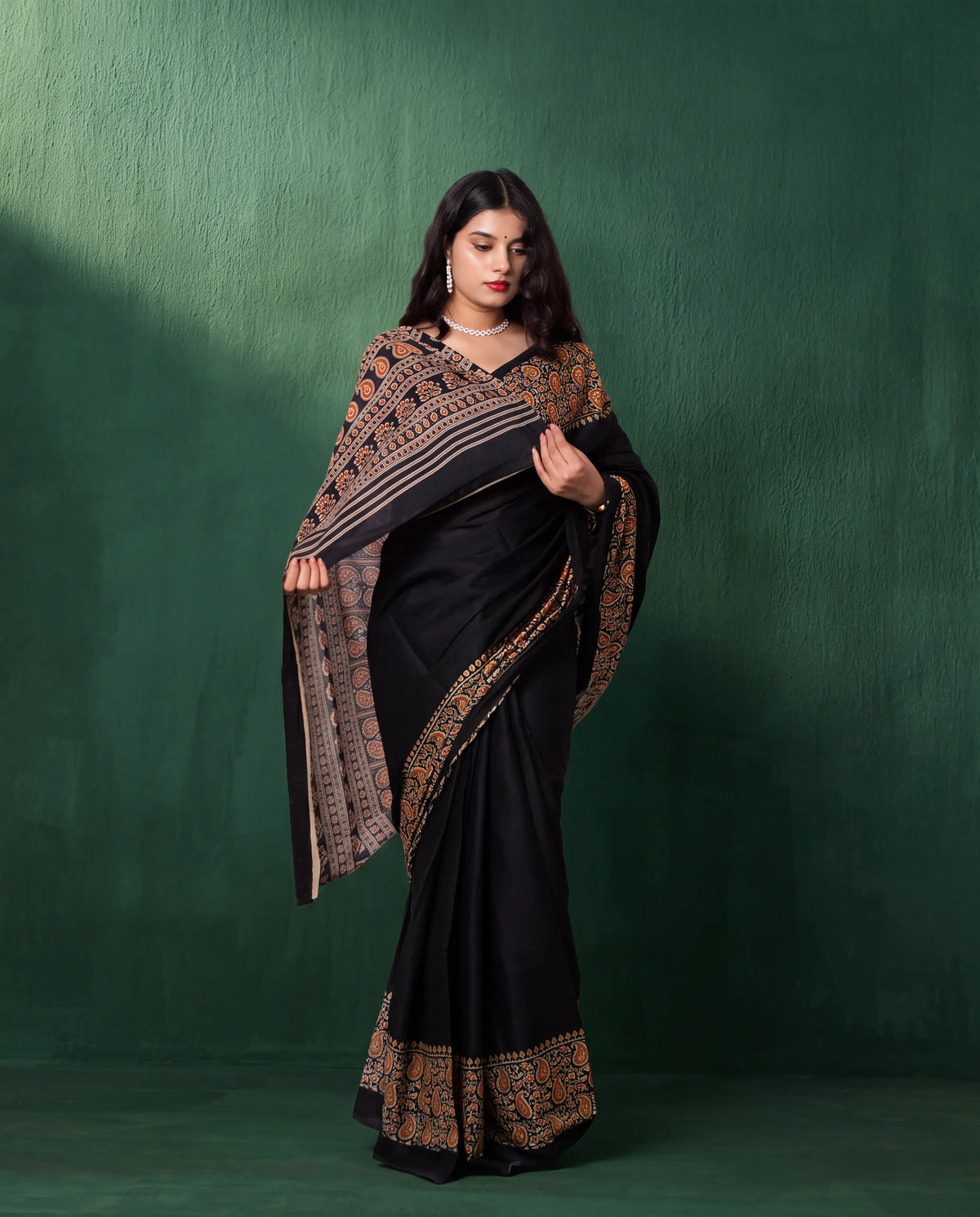 A woman modeling a black sari with ornate gold and brown border patterns, standing against a textured dark green studio backdrop.