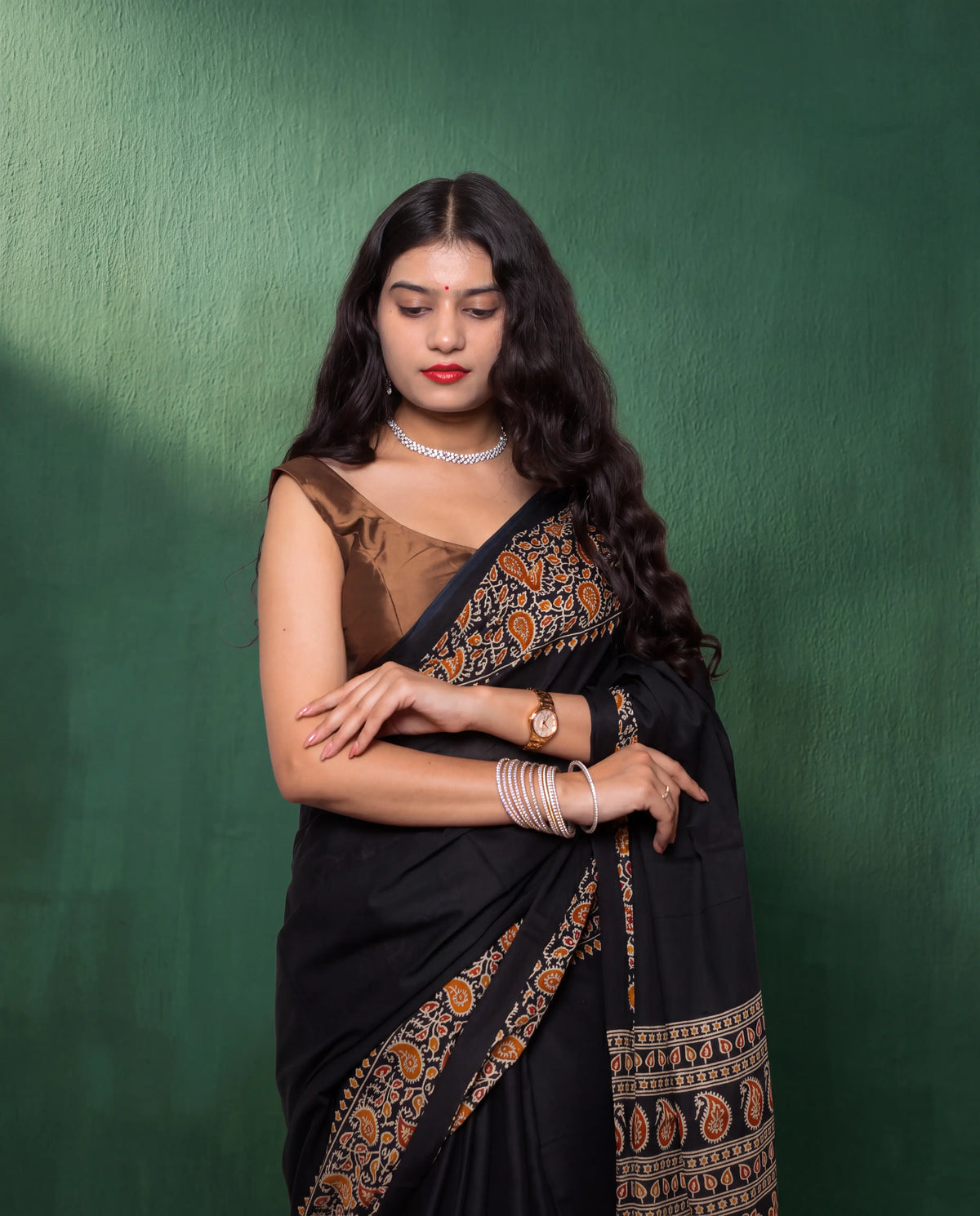 A woman modeling a black saree with intricate gold and orange paisley border patterns, paired with a brown sleeveless blouse, standing against a textured dark green backdrop.