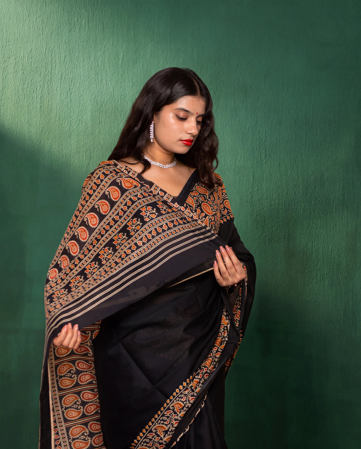 A woman wearing a black saree with an ornate peach, gold, and beige border, posed against a textured dark green wall.